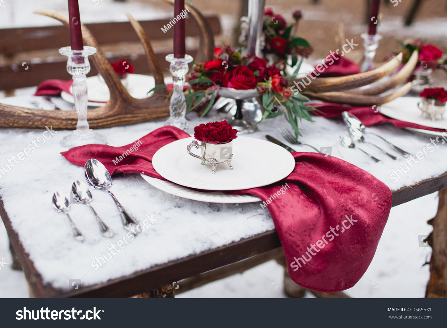 Wedding table setting in marsala colors with plates  cutlery  red floral compositions  candles  velvet napkins  deer horns on table covered with snow