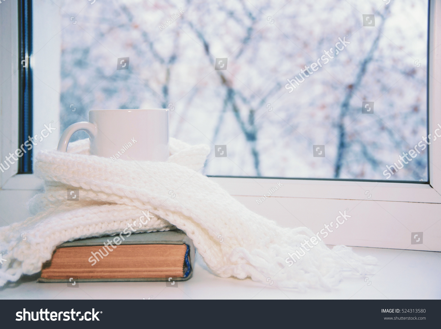 Mug of hot tea and warm woolen knitting on windowsill against snow landscape from outside.