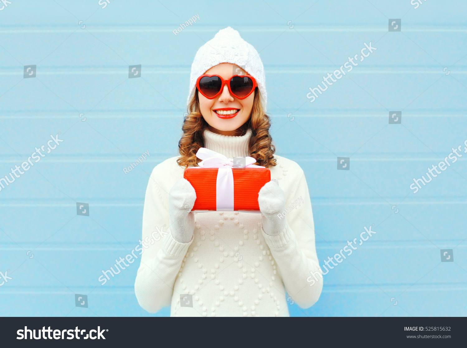 Christmas happy smiling young woman with gift box wearing a knitted hat sweater sunglasses over blue background