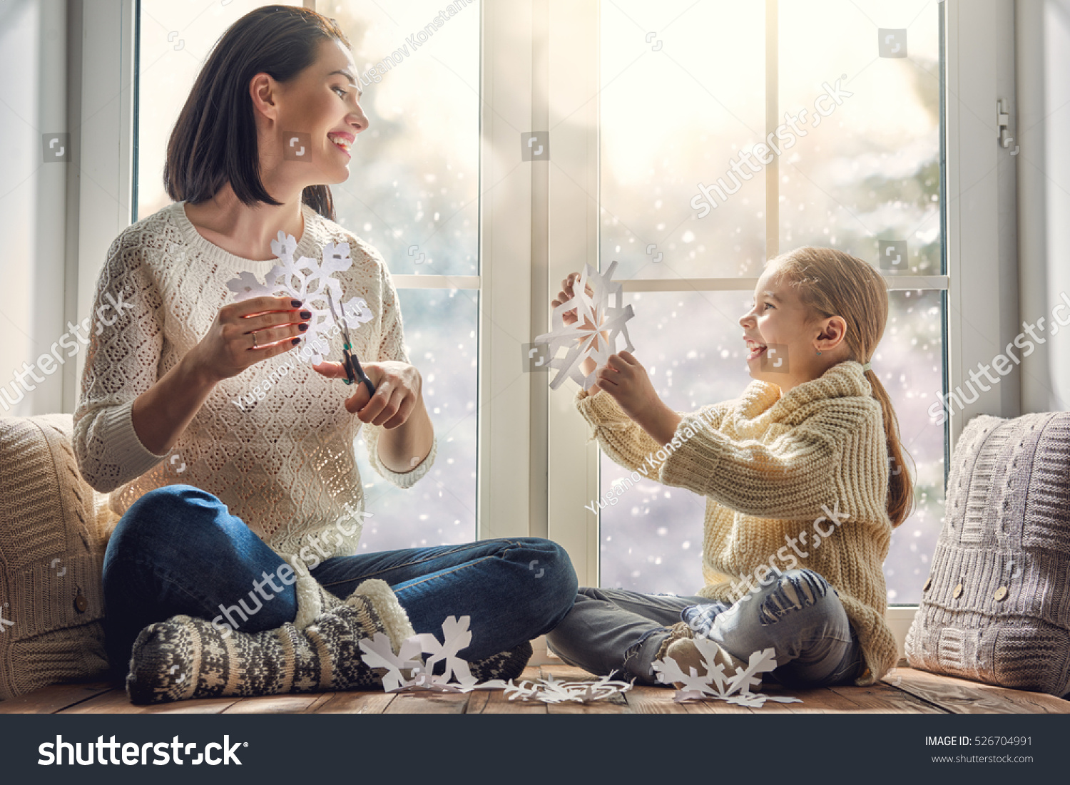 Merry Christmas and happy holidays! Happy loving family sitting by the window and making paper snowflakes for decoration windows. Mother and child creating decorations.