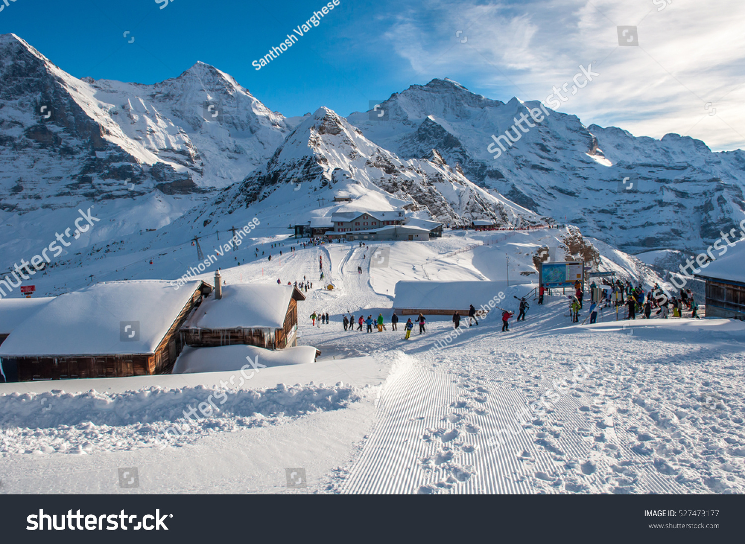 Lots of people with snowboards and skis on the mountain top getting ready for snow fun. Sun shining/sunlight on the snow covered wooden cottages  in Engelberg  Mount Titlis  Switzerland.  