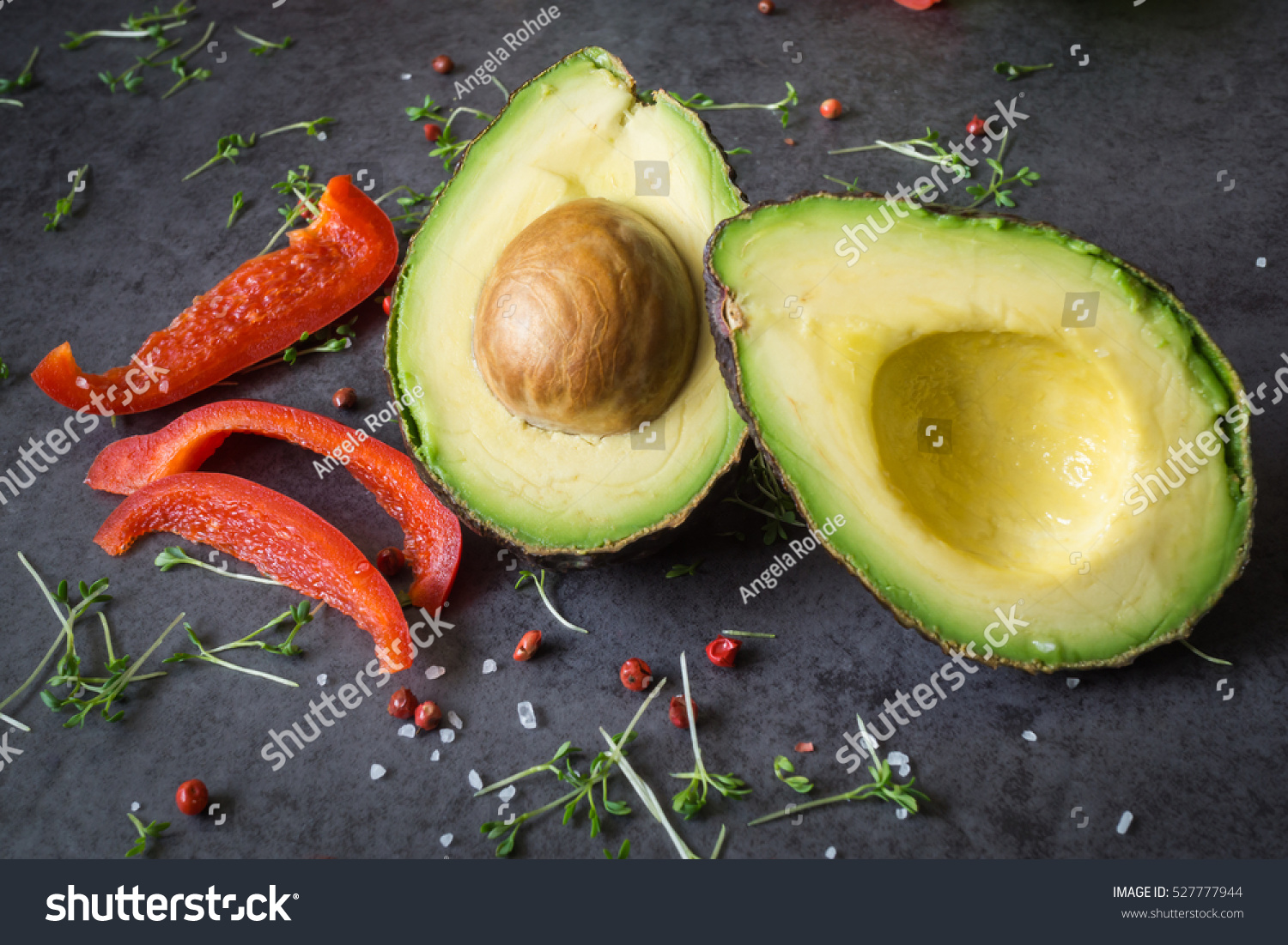 A halved avocado with kernel  sliced ??paprika  coarse sea salt  cress and dried red berries  background black slate