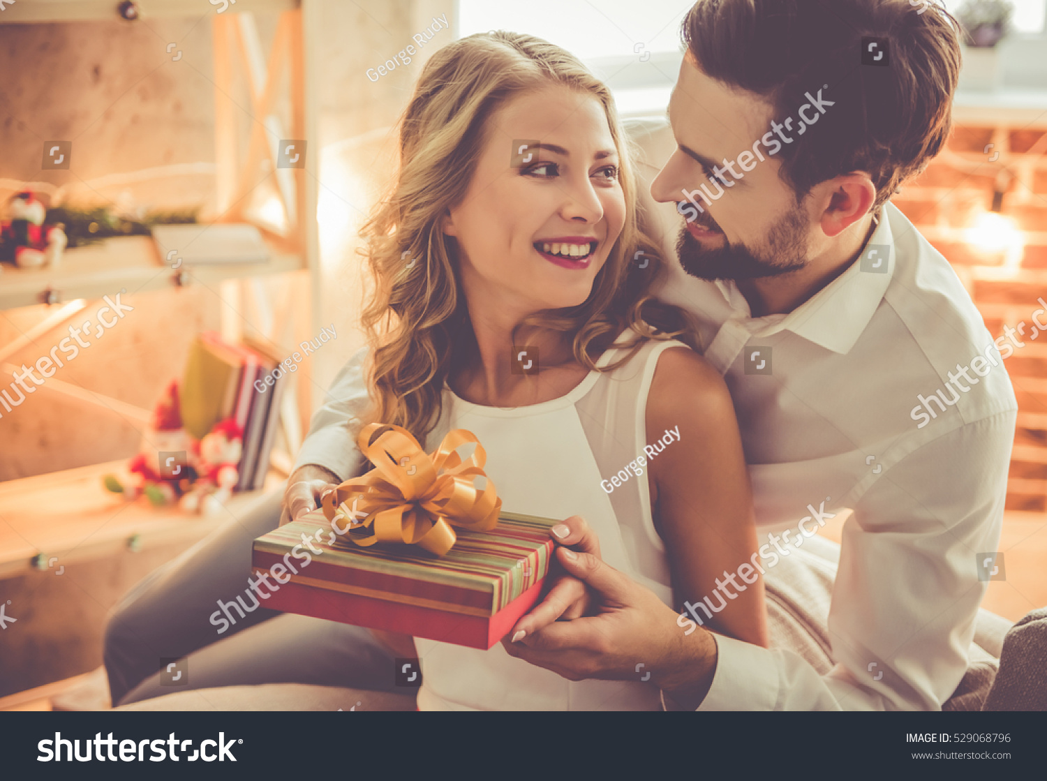 Beautiful young couple is celebrating at home. Handsome man is giving his girlfriend a gift box