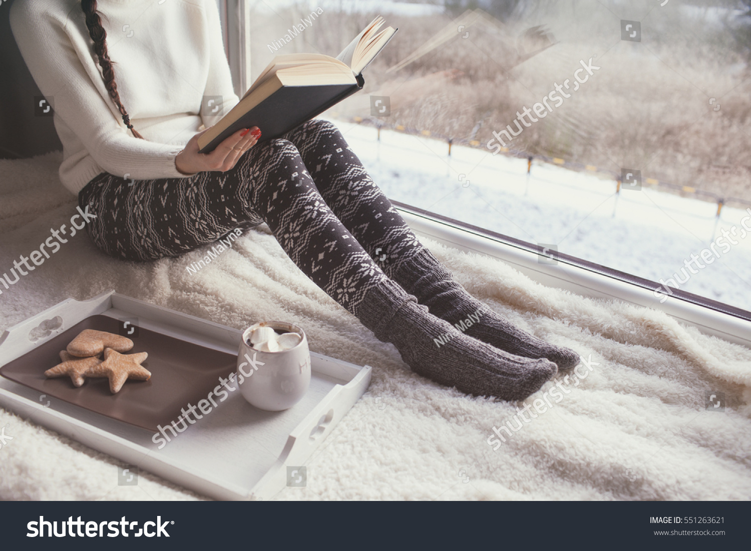 Young woman with cup of coffee and gingerbread is reading a book sitting home relaxing by the window. Snow winter home chill out concept