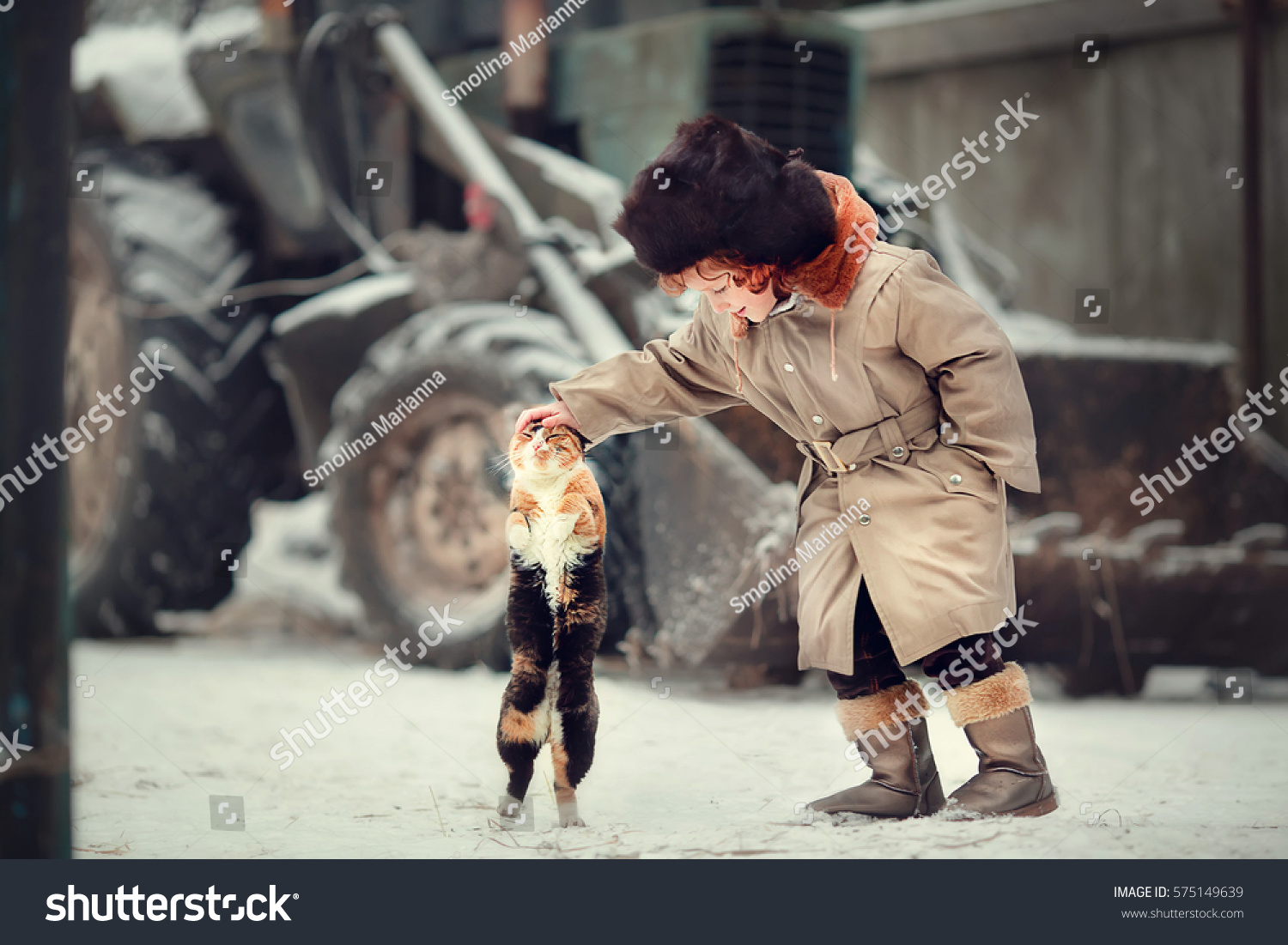 Funny little red-haired boy in earflaps hat is stroking a cat in the country in winter against a tractor background. Cat is standing on hint legs. Image with selective focus and toning.