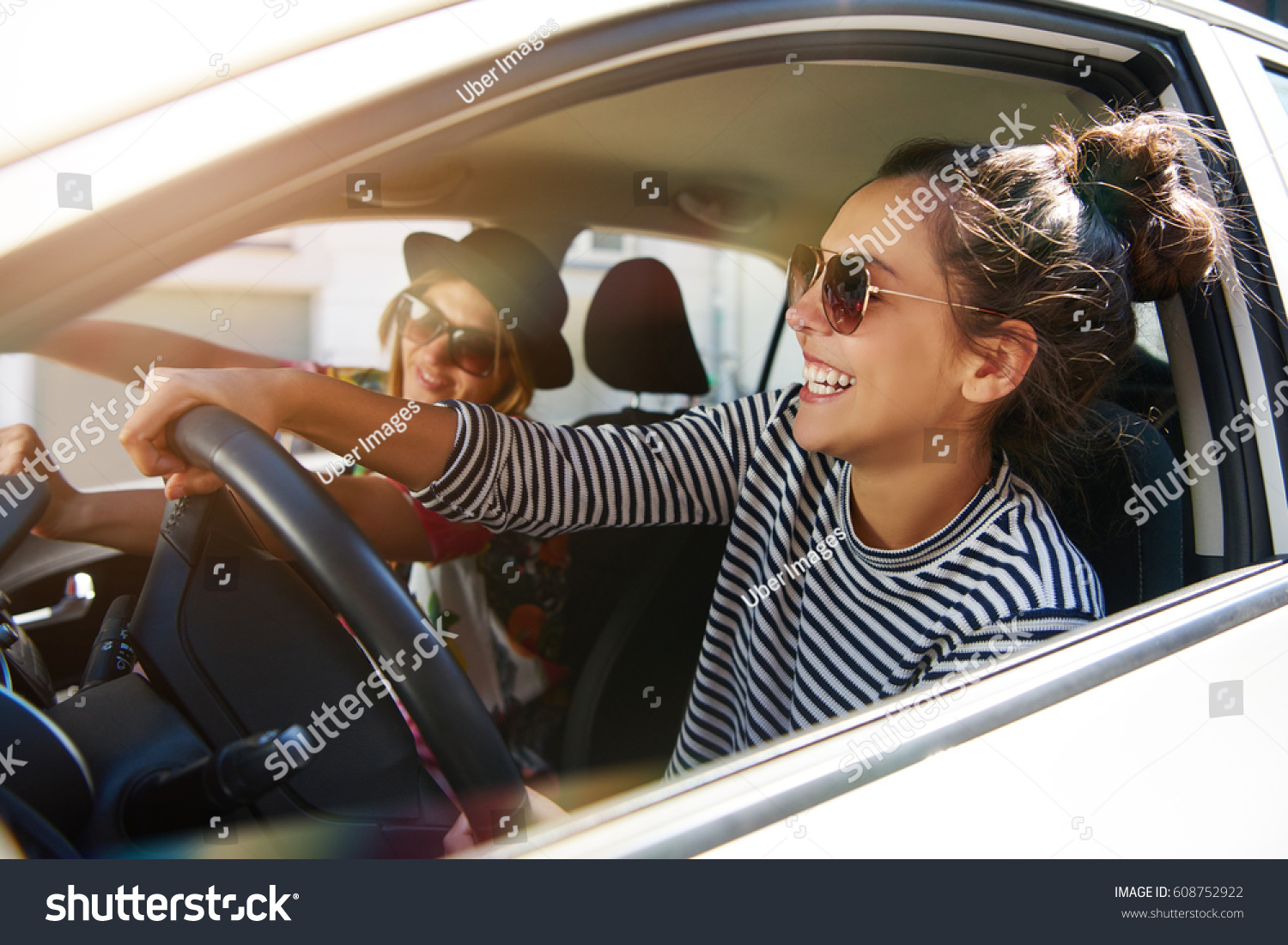 Two fun young women in sunglasses driving in a car in town laughing and smiling as they socialise together view through open side window