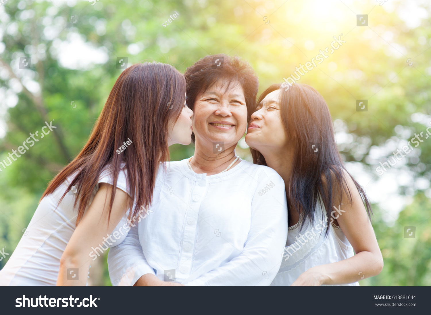 Portrait of Asian daughters kissing elderly mother senior adult woman and grown child. Outdoors family at nature park with beautiful sun flare.