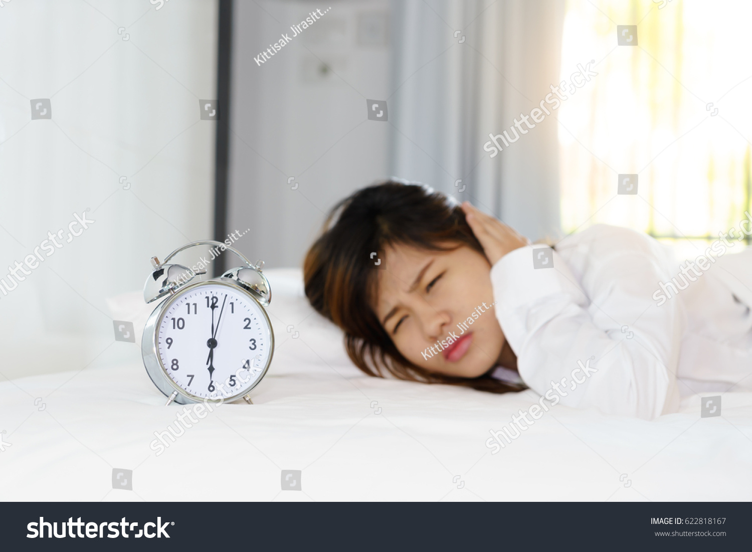 Sleepy young woman looking at alarm clock with hands covering her ears in the morning. early wake up