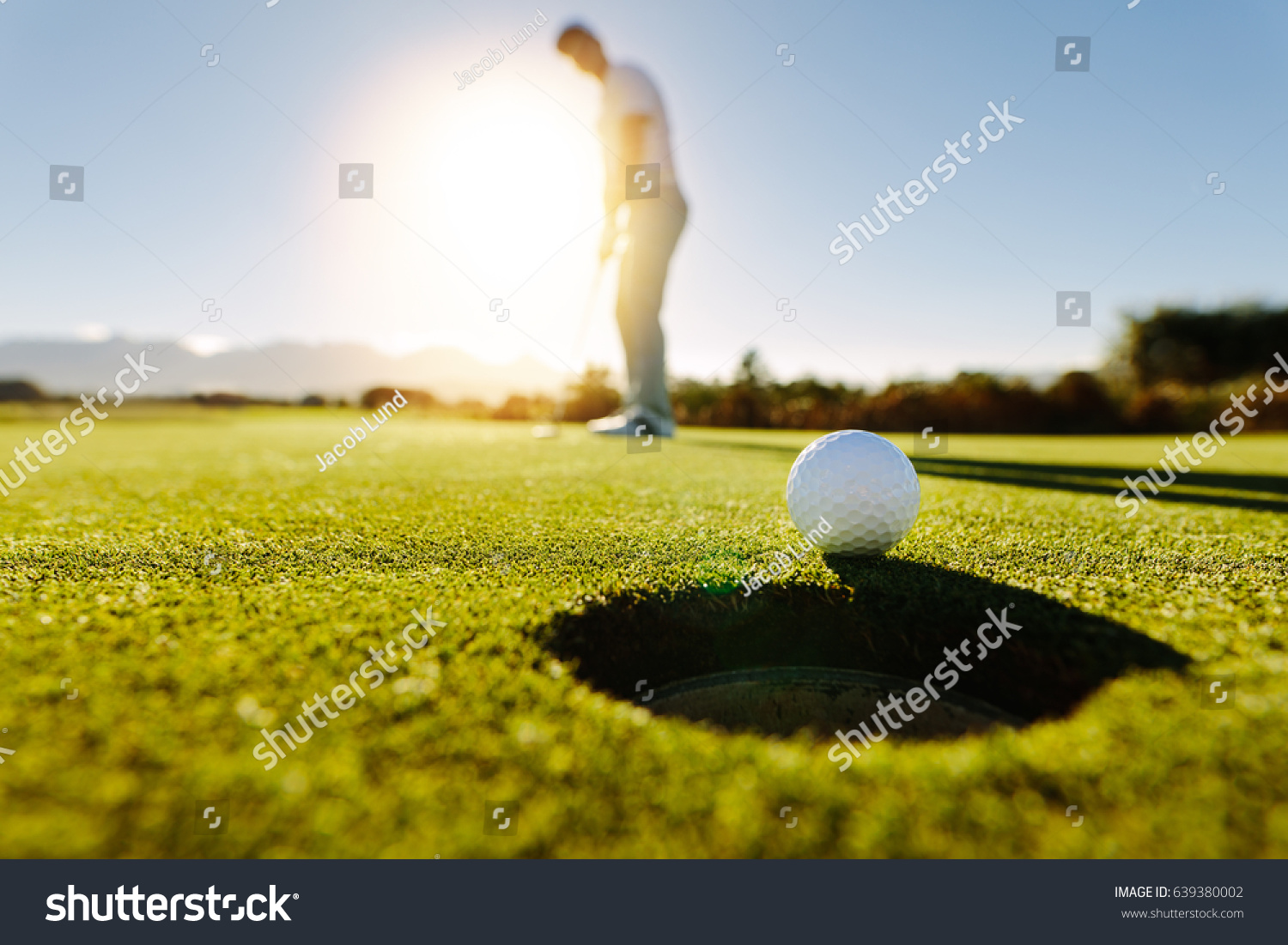 Pro golfer putting golf ball in to the hole. Golf ball by the hole with player in background on a sunny day.