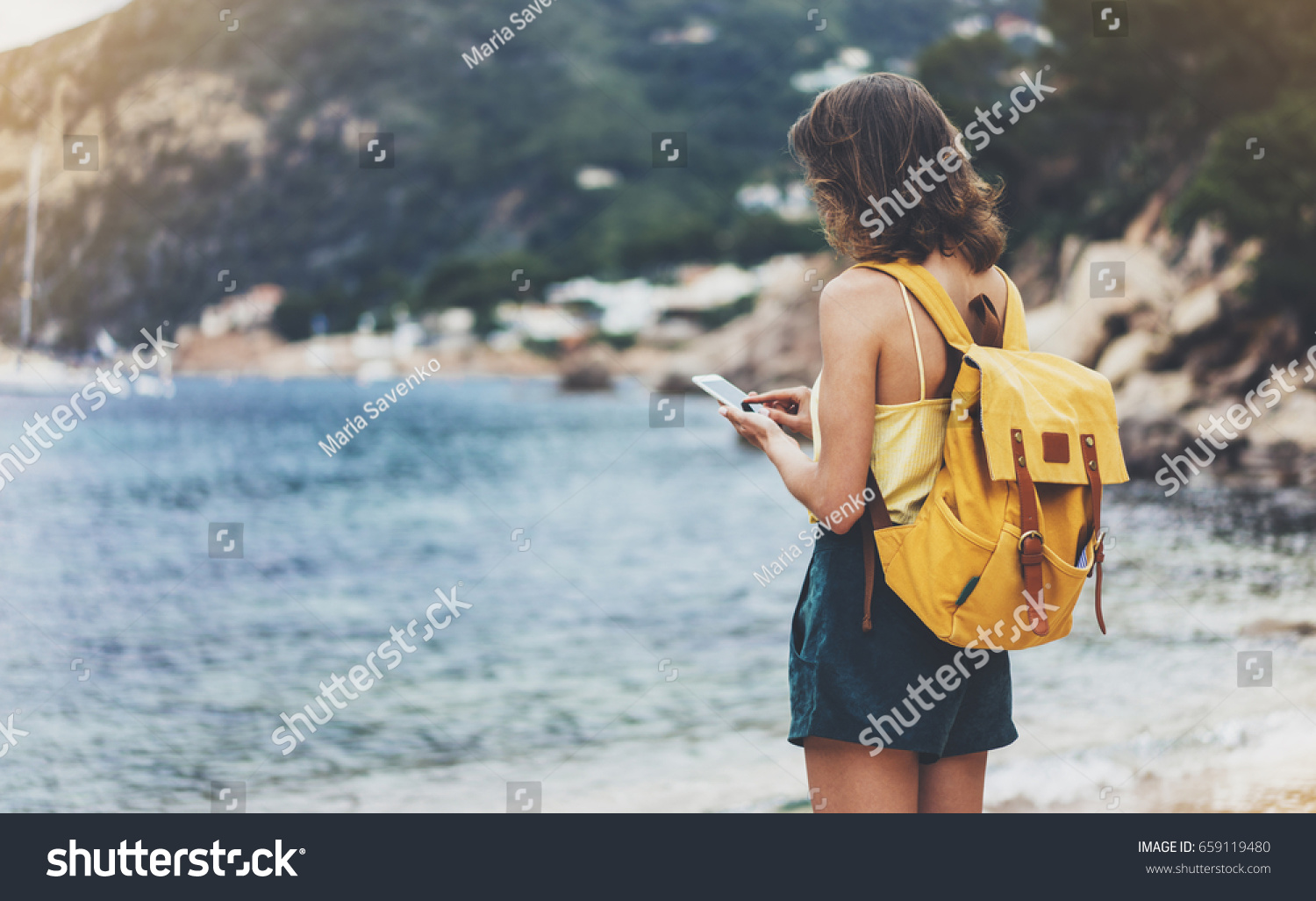 Hipster girl with backpack hold on smart phone gadget in sand coastline. Traveler using in female hand mobile on background beach seascape horizon. Tourist look on blue sun ocean summer lifestyle