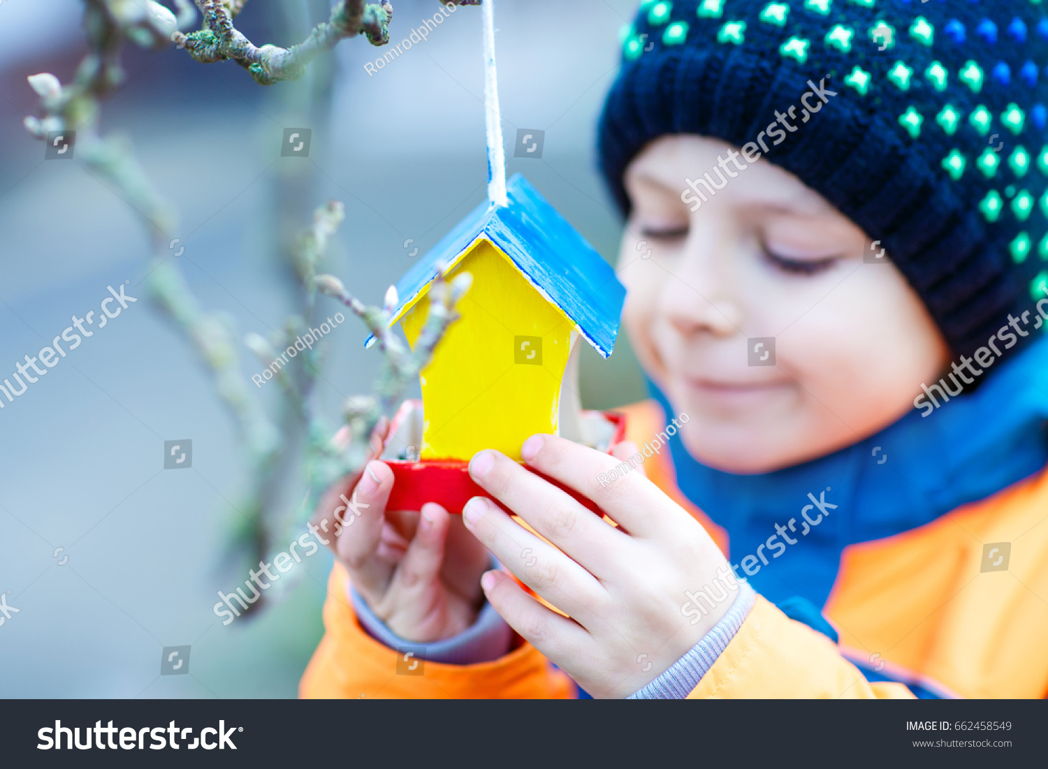 Little kid feeding birds in winter. Child hanging colorful selfmade bird house on tree on frosty cold day. Toddler in colorful wam clothes. Selective focus on hands and feeder with seeds