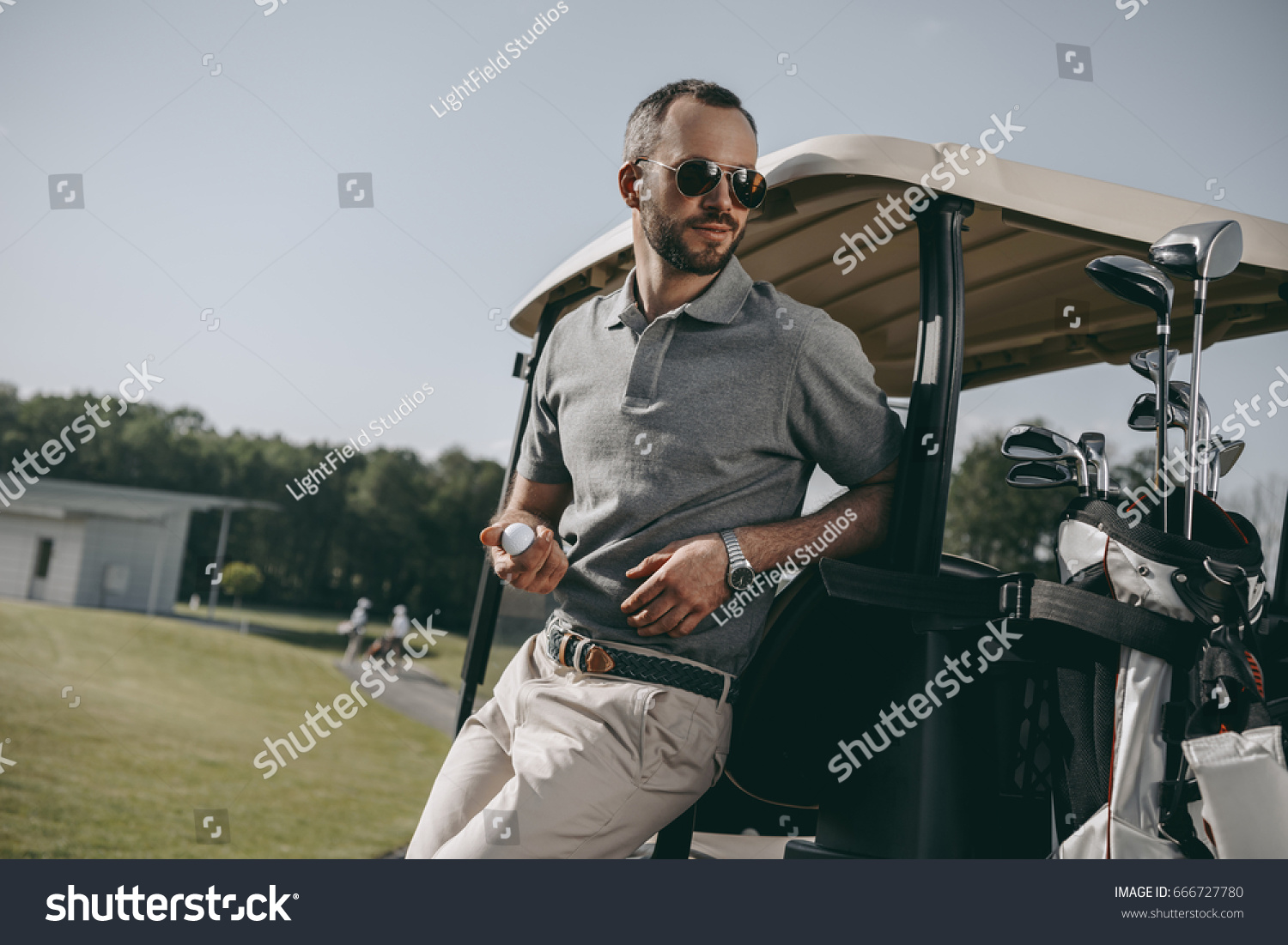 stylish golfer holding golf ball and looking away while leaning on golf cart