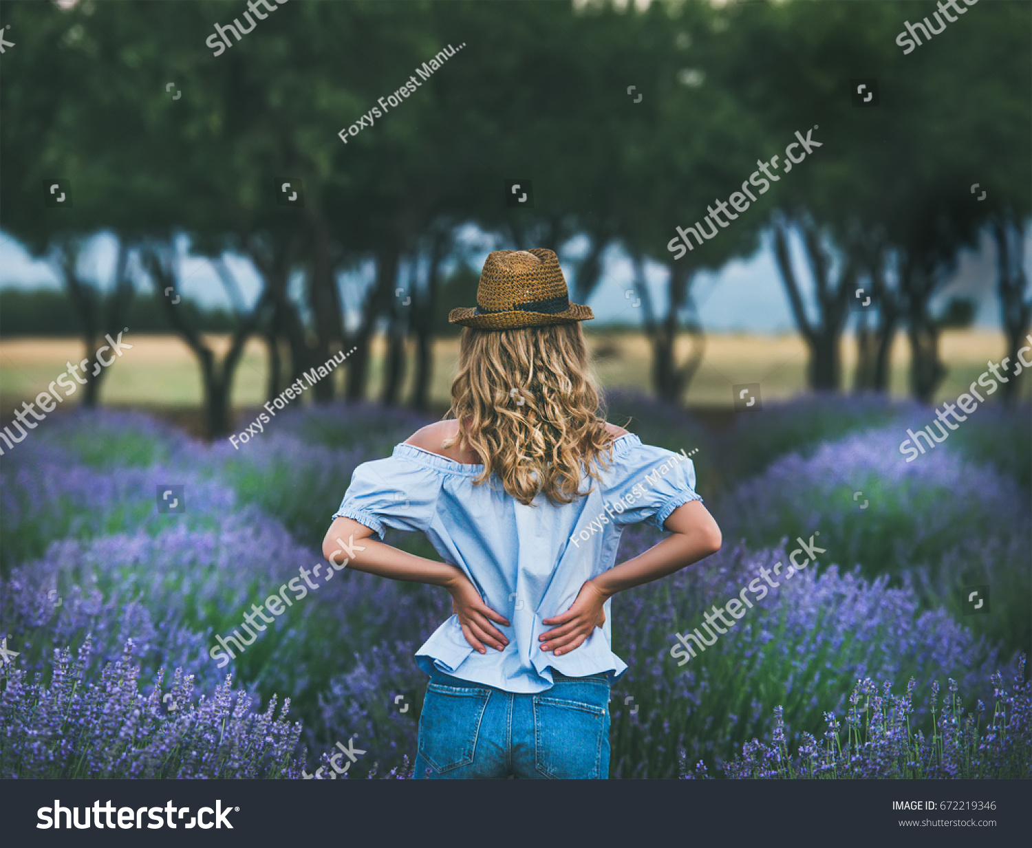 Young blond woman traveller wearing straw hat denim shorts and blue off-shoulder blouse standing in lavender field surrounded with lavender flowers in Isparta region of Turkey