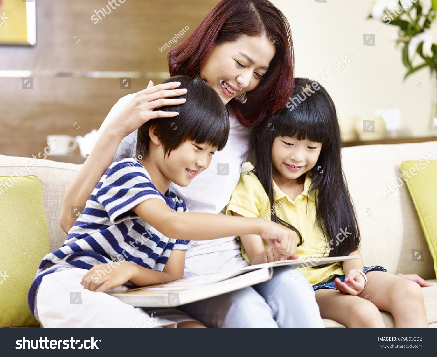 young asian mother and children sitting on couch at home reading a book together happy and smiling.