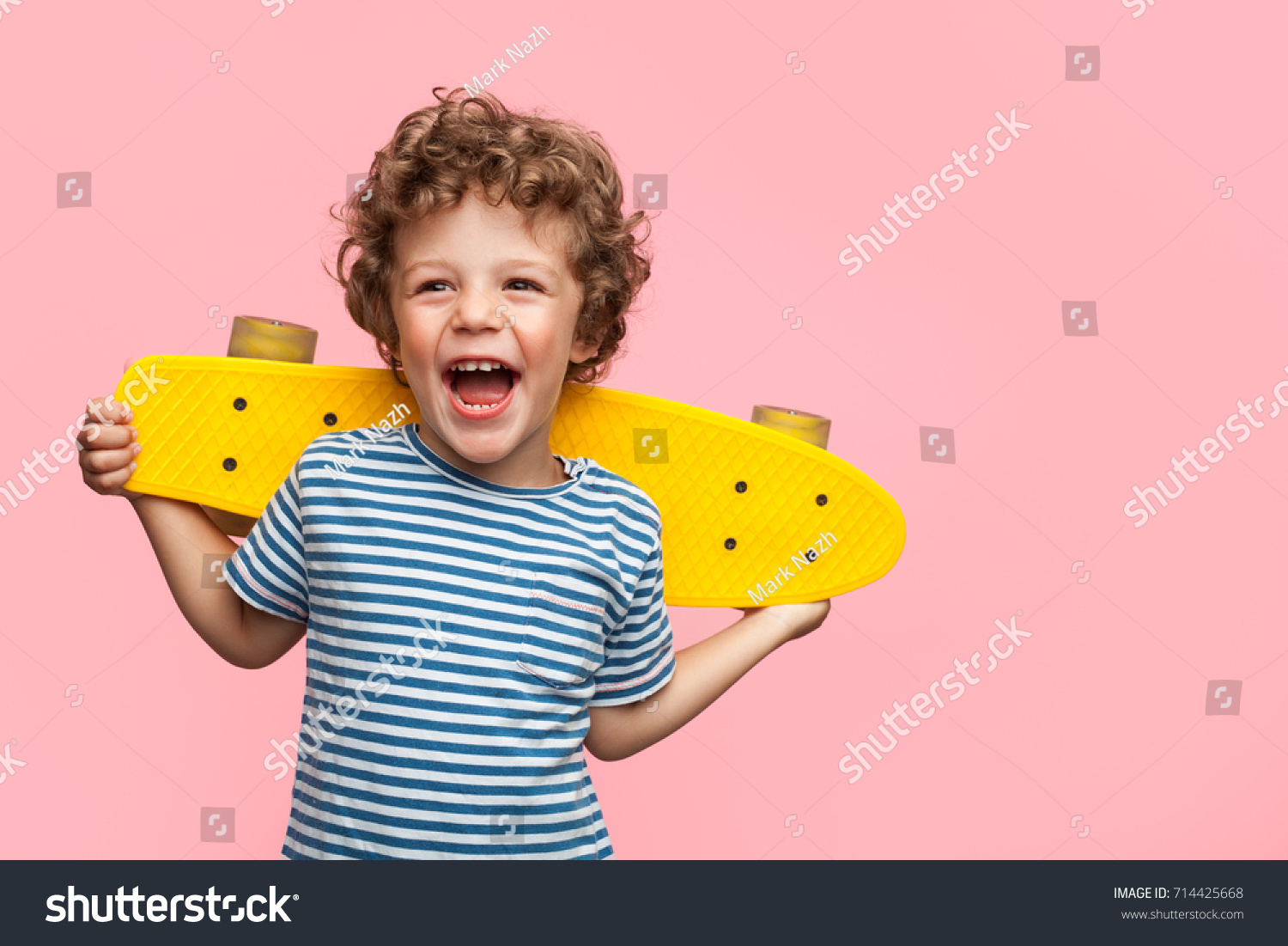Charming curly boy holding yellow longboard and looking away on pink background. 