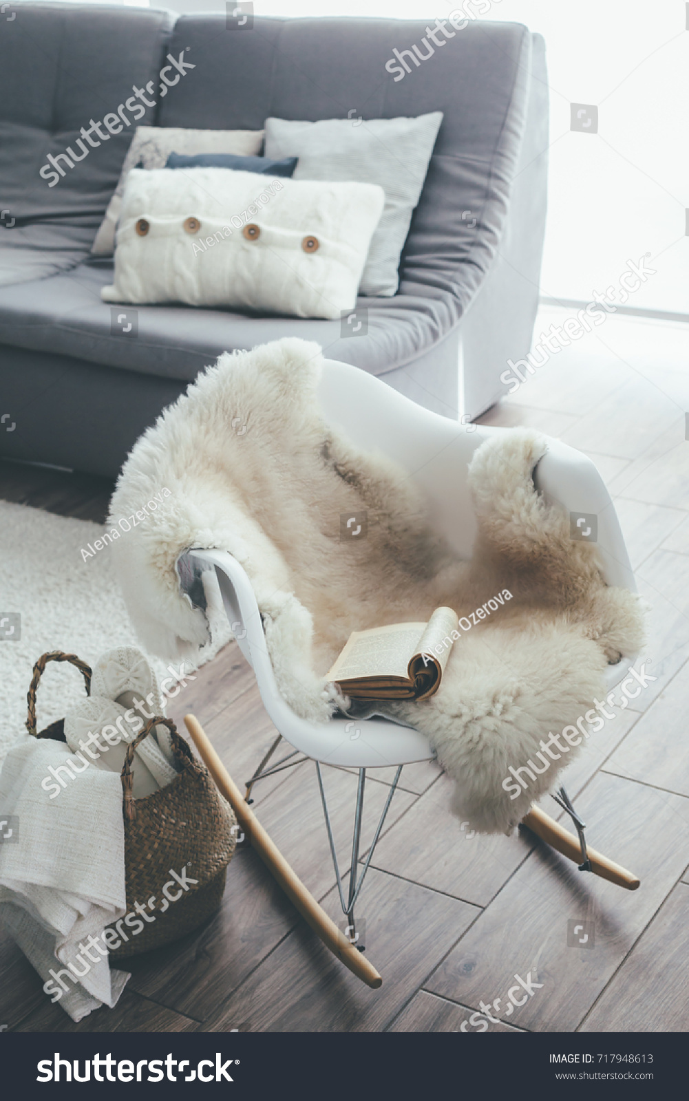 Still life details of living room. Sheep skin rug on modern armchair by the sofa with cushions. Reading book on the rocket chair. Cozy winter scene in Scandinavian interior.