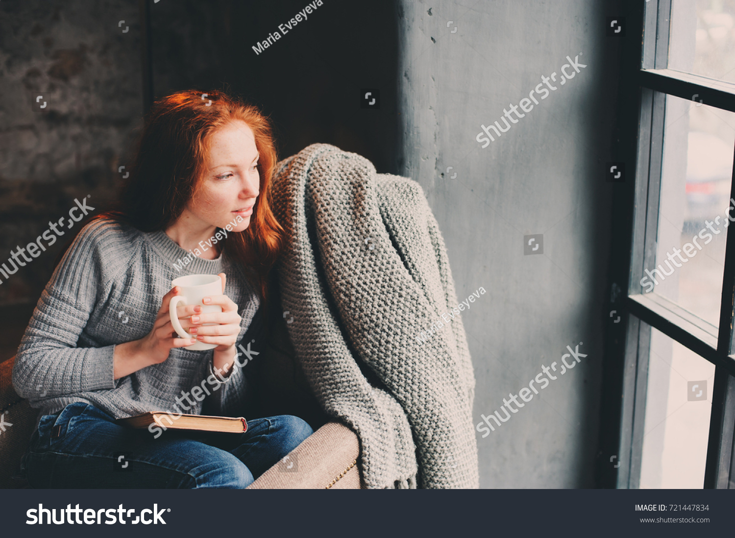 happy redhead woman relaxing at home in cozy winter or autumn weekend with book and cup of hot tea sitting in soft chair with blanket