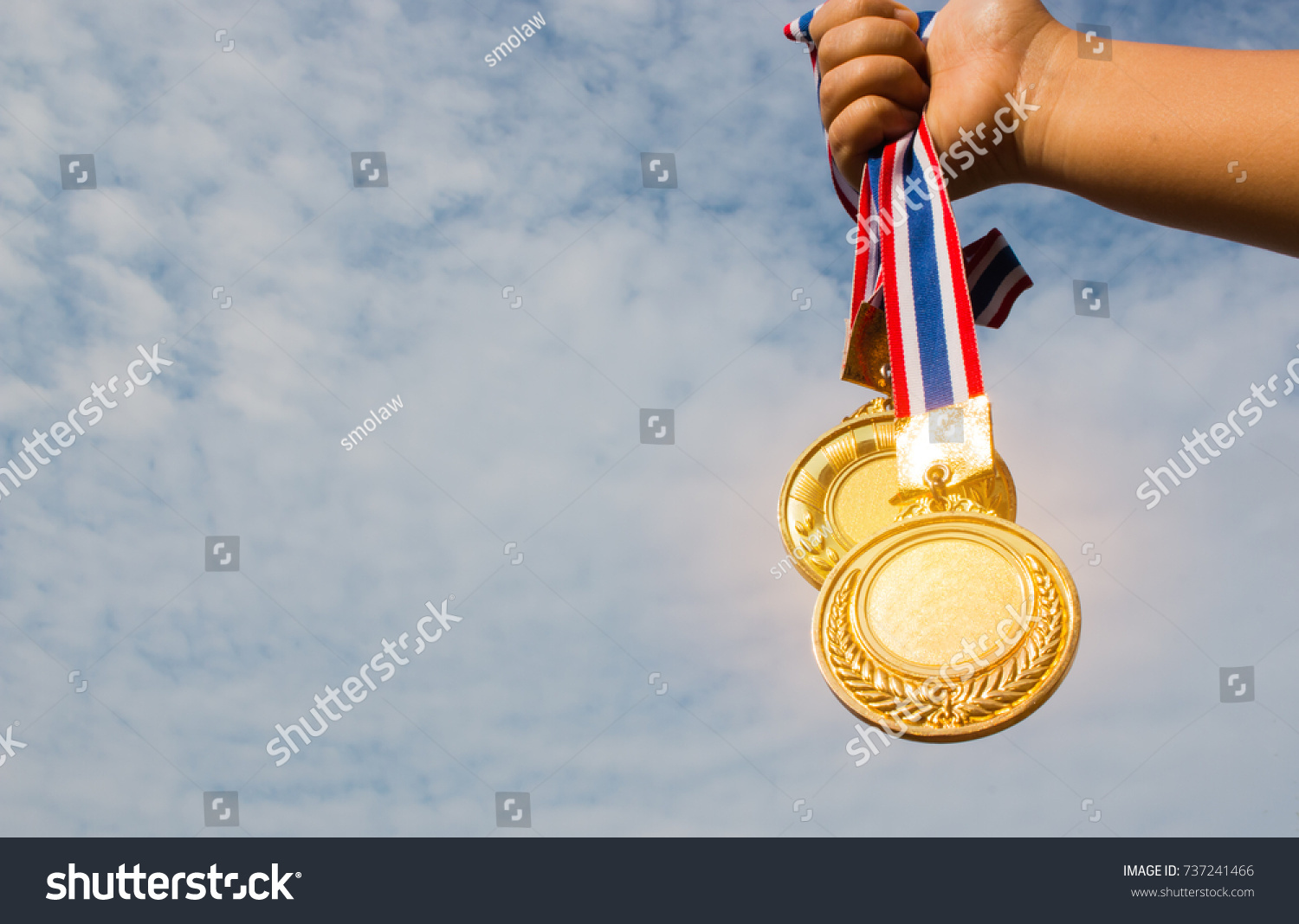 Winner hand raised and holding two gold medals with Thai ribbon against blue sky. Golden medal is medal awarded for highest achievement for sport or business. Success Awards concept