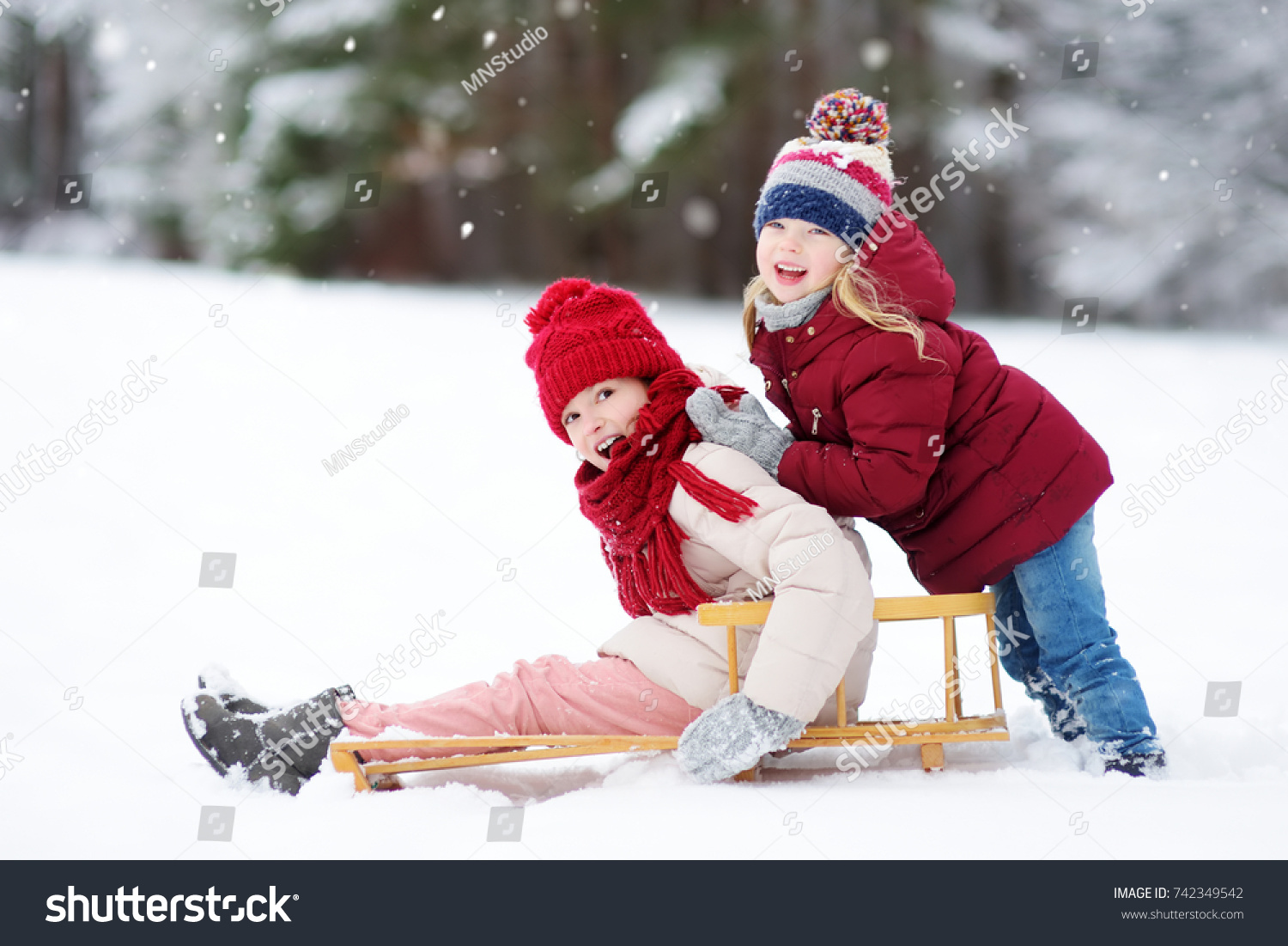 Two funny little girls having fun with a sleight in beautiful winter park. Cute children playing in a snow. Winter activities for kids.
