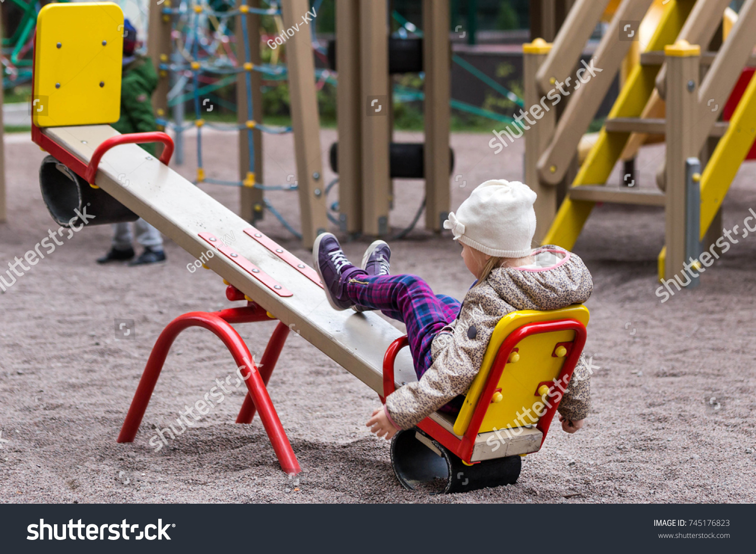cute caucasian girl in jacket and hat sitting alone on a seesaw
