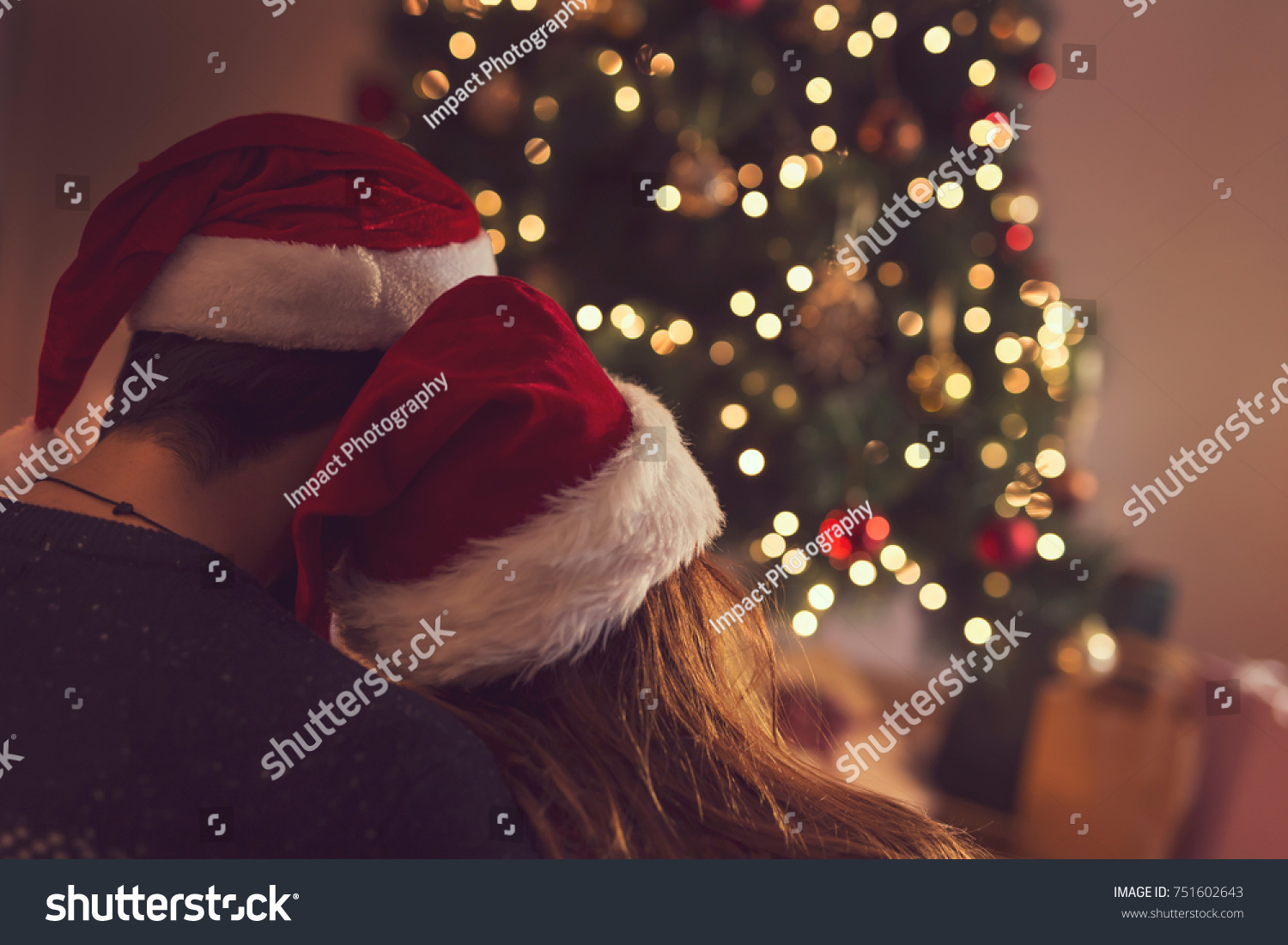 Couple in love sitting next to a Christmas tree wearing Santa's hats hugging and looking away from the camera towards the tree. Selective focus on the girl's hat