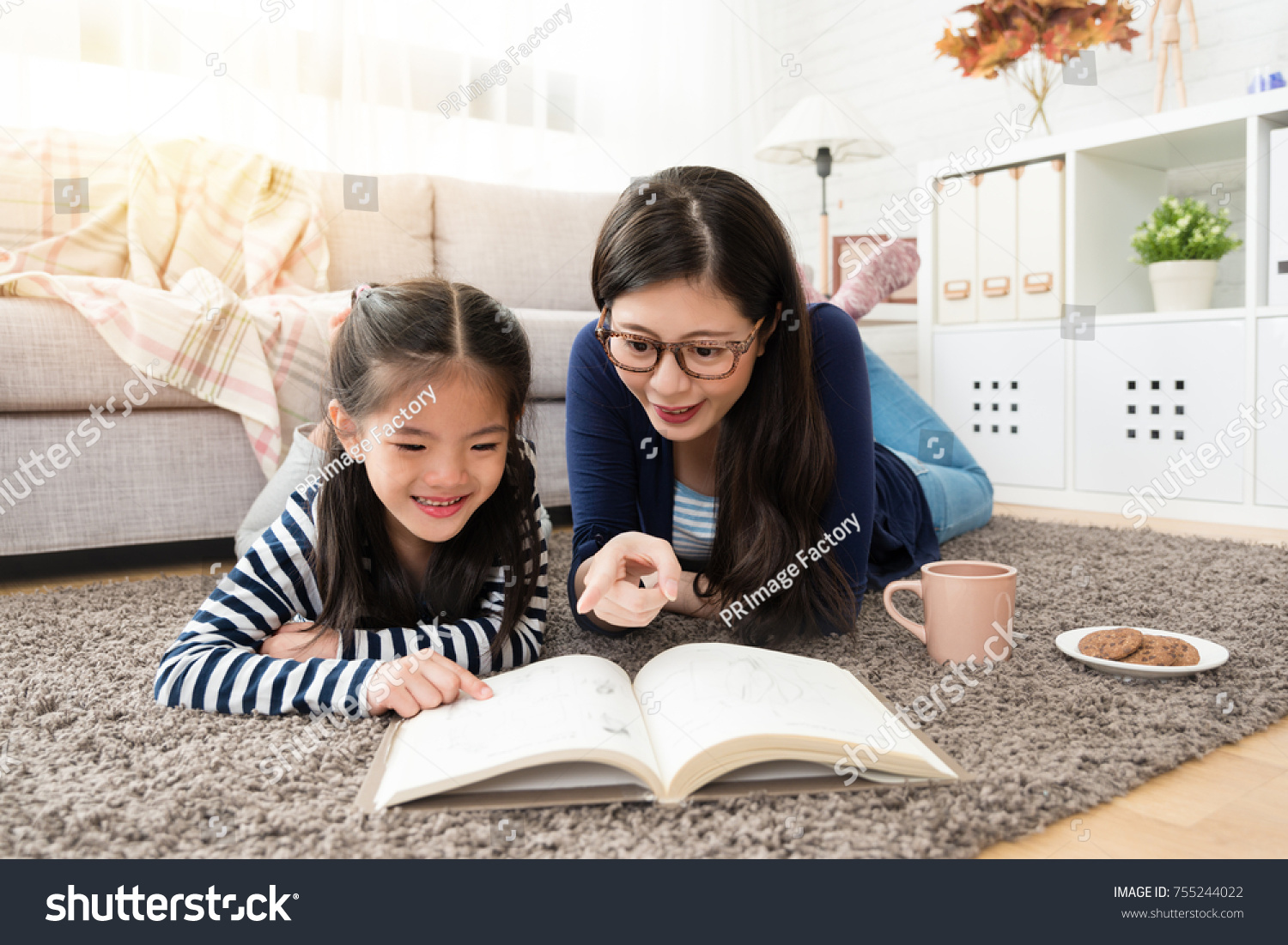 charming Asian mother and her daughter reading book and enjoy hot chocolate and coffee sitting on floor of the living room at home