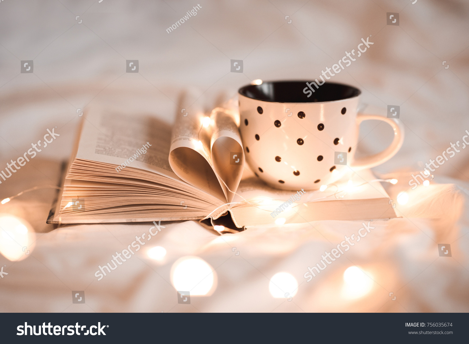 Open book with folded sheets in heart shape and cup of tea in bed with Christmas lights closeup. Good morning. Breakfast time.