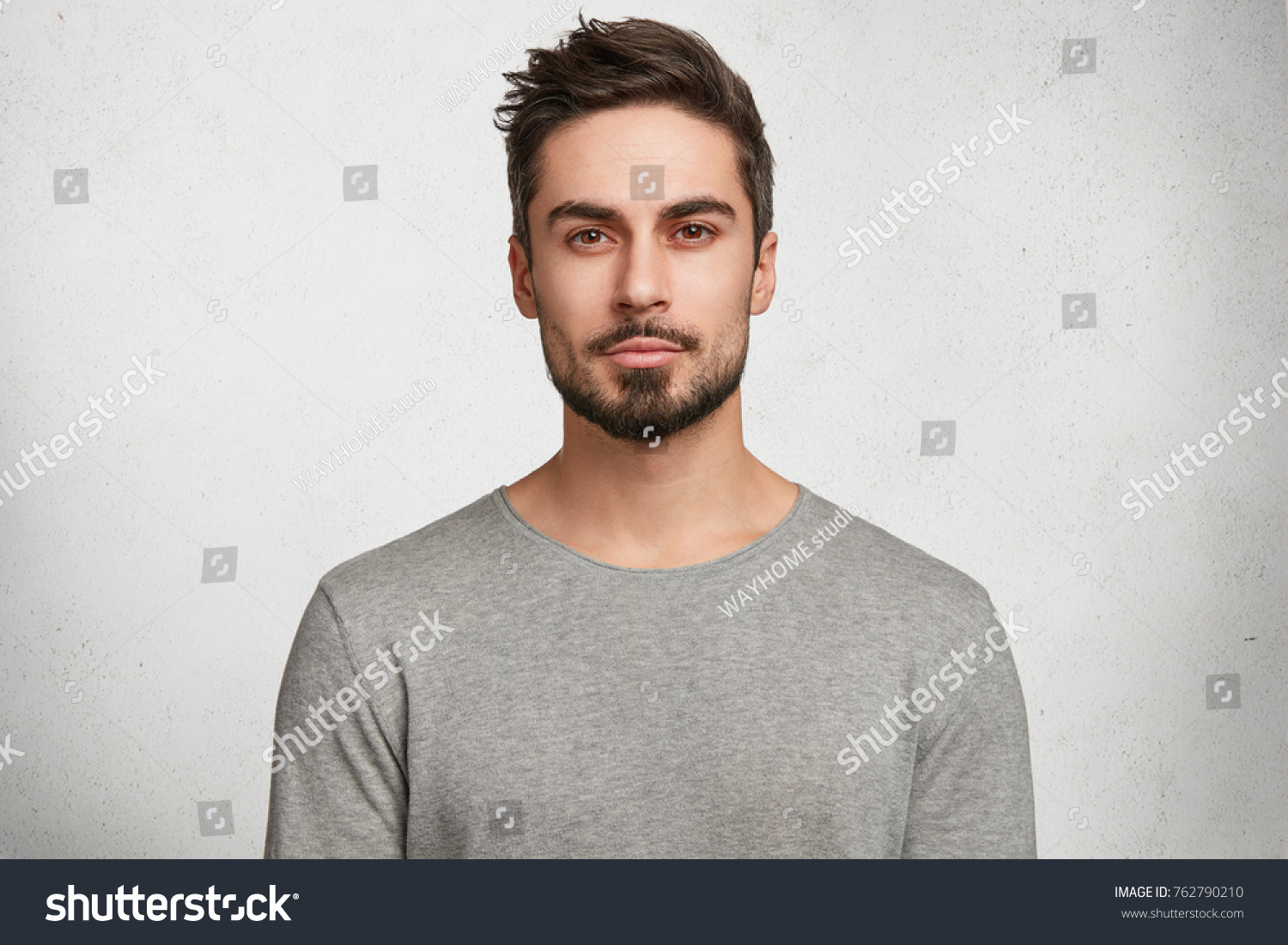 Isolated shot of young handsome male with beard  mustache and trendy hairdo  wears casual grey sweater  has serious expression as listens to interlocutor  poses in studio against white background