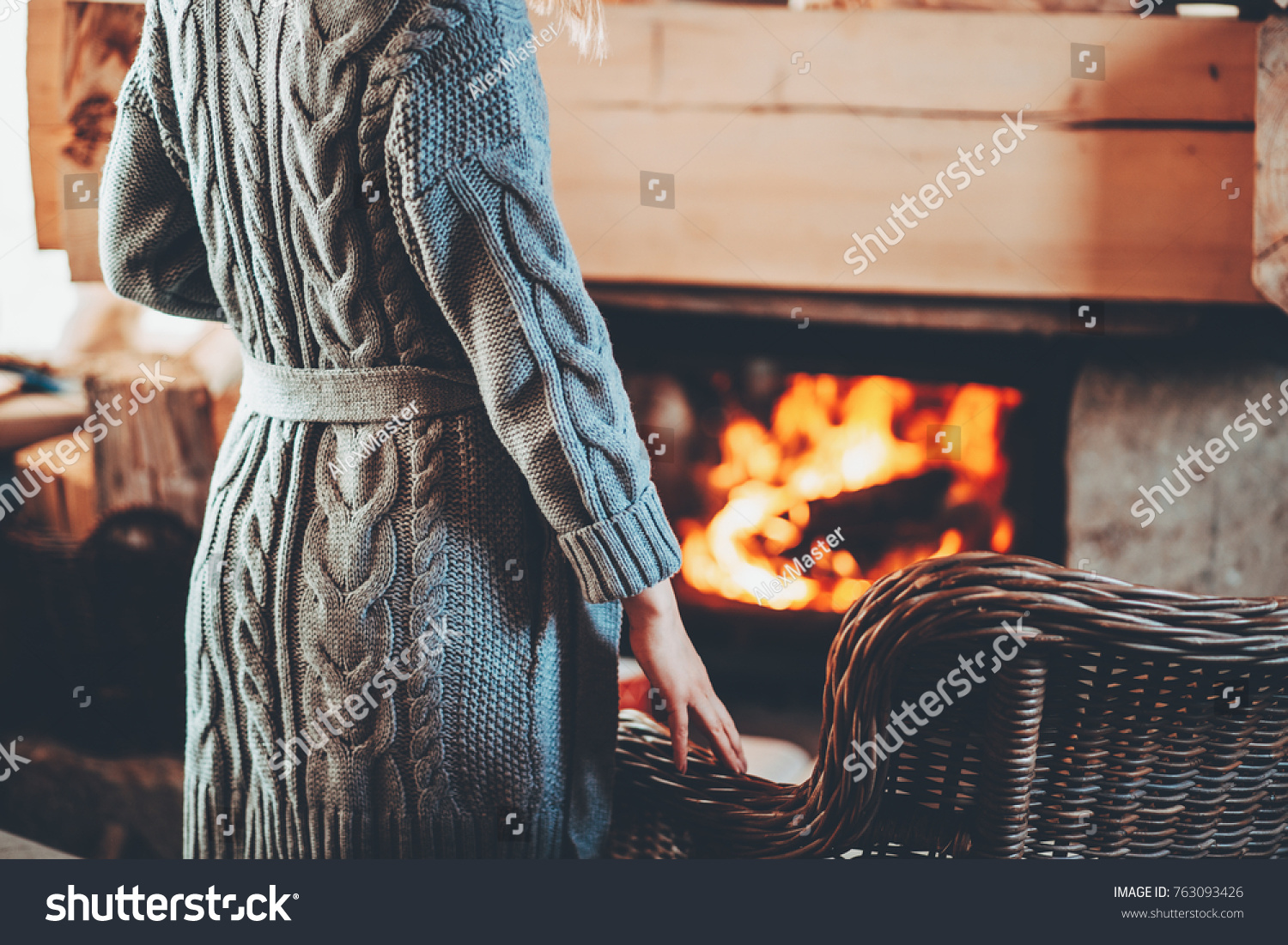 Woman in warm knitted cardigan sweater by fireplace. Woman relaxes by warm fire with a cup of hot drink and warming up her feet in woollen socks. Cozy atmosphere. Winter and Christmas holidays concept
