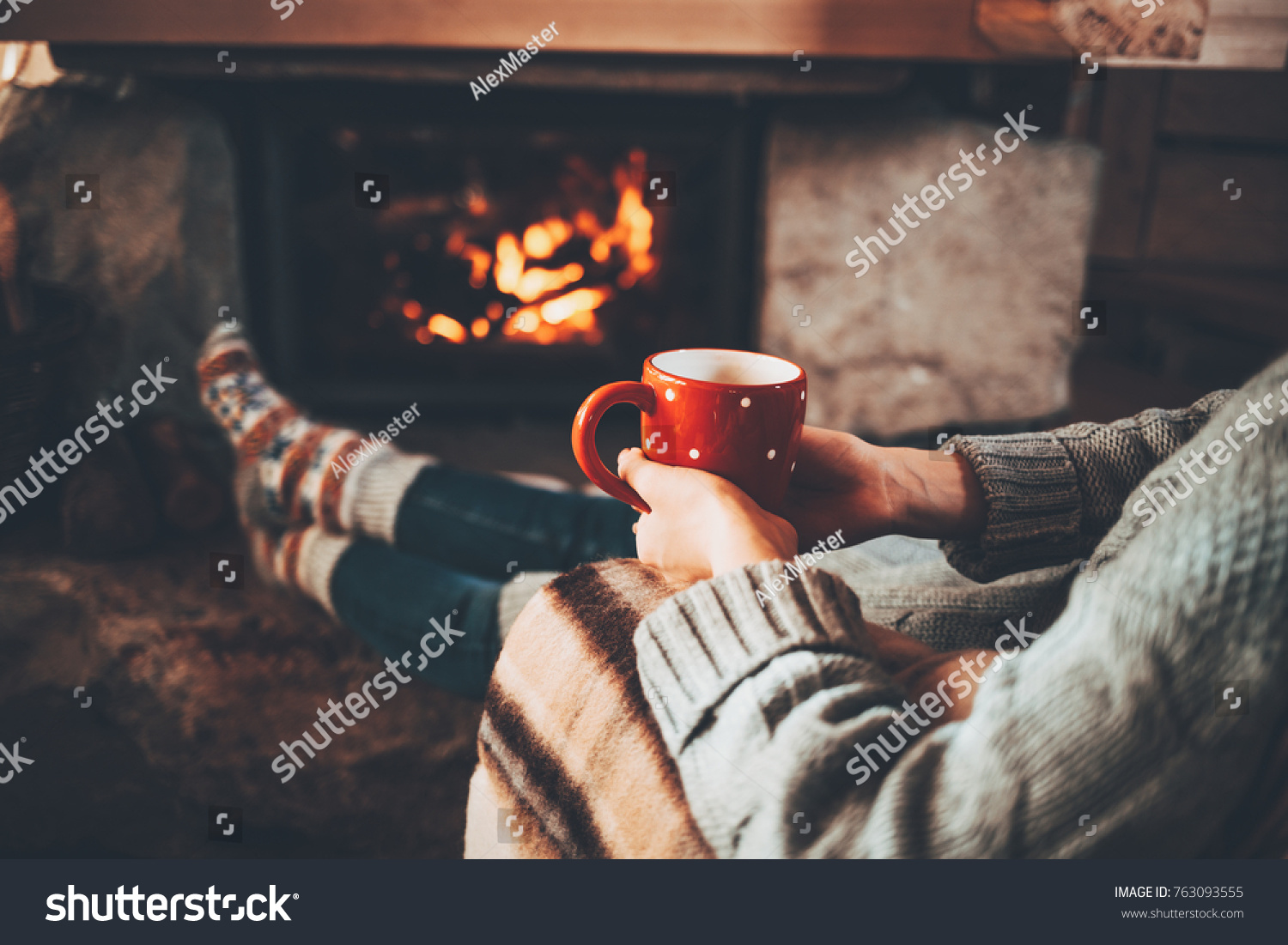 Feet in woollen socks by the Christmas fireplace. Woman relaxes by warm fire with a cup of hot drink and warming up her feet in woollen socks. Cozy atmosphere. Winter and Christmas holidays concept.