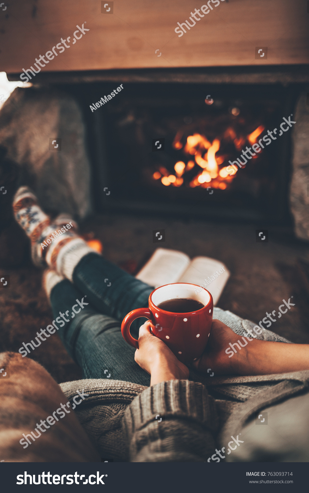 Feet in woollen socks by the Christmas fireplace. Woman relaxes by warm fire with a cup of hot drink and warming up her feet in woollen socks. Cozy atmosphere. Winter and Christmas holidays concept.