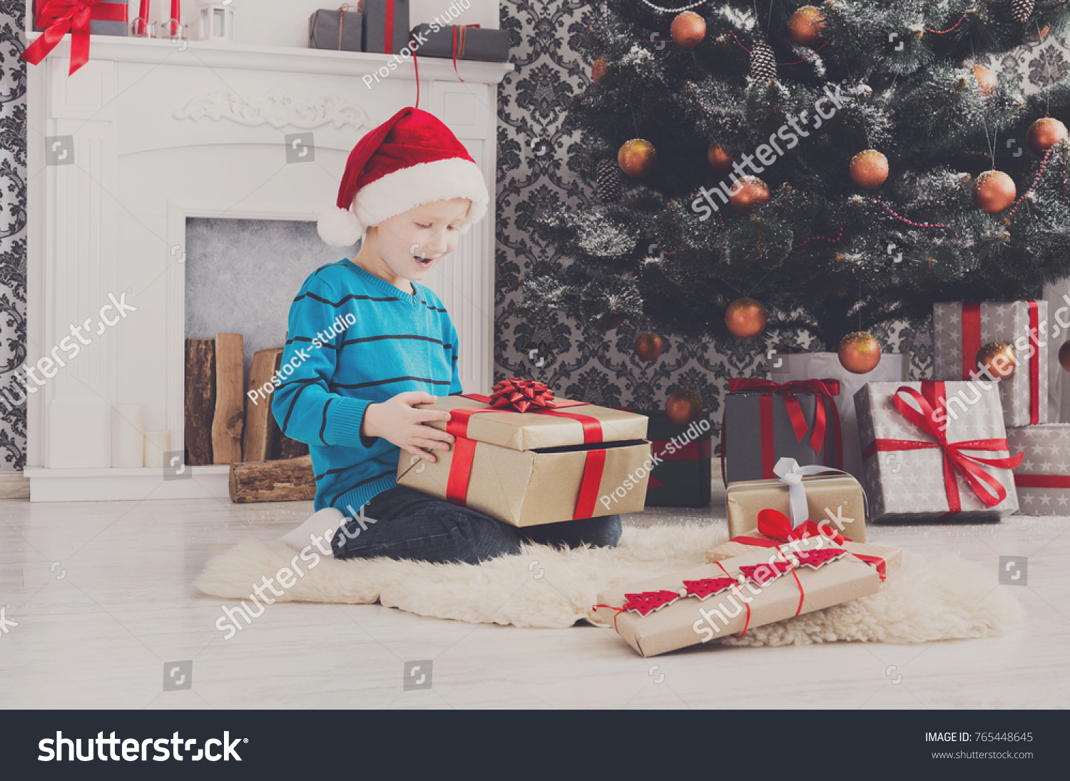 Cute happy boy in santa hat unwrap christmas present box on holiday morning in beautiful room interior. Male child open Xmas gifts near big decorated fir tree and fireplace. Winter holidays concept