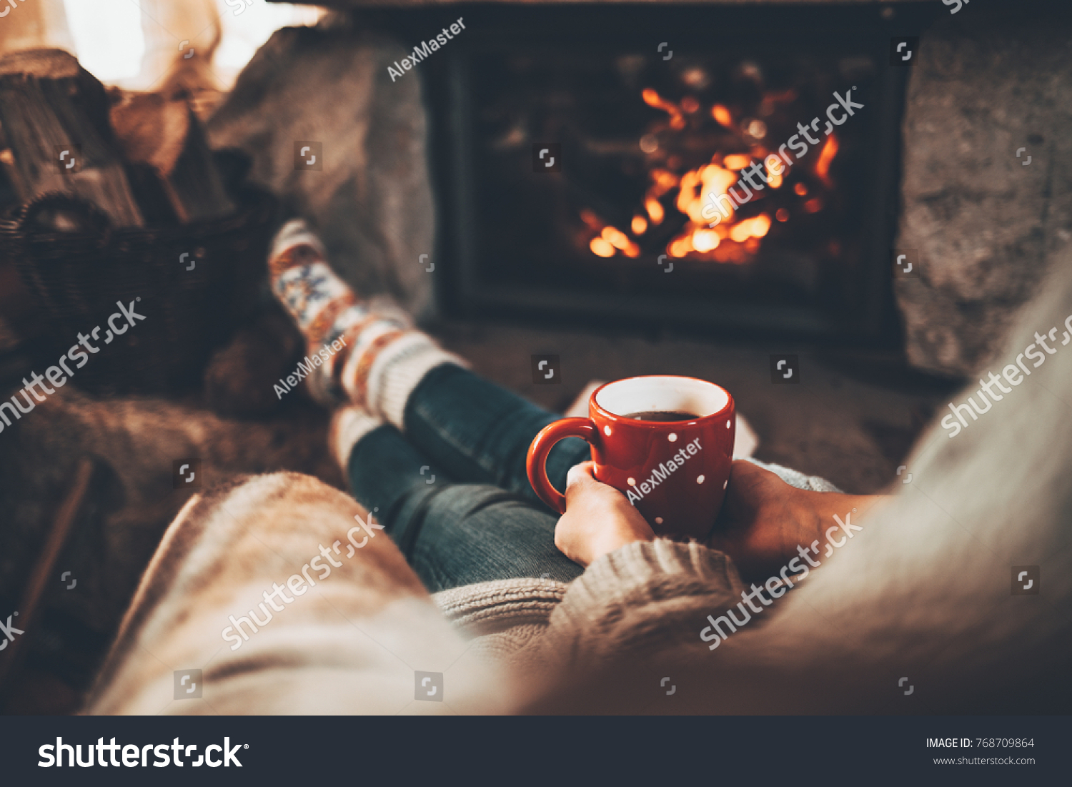 Woman in warm knitted cardigan sweater by fireplace. Woman relaxes by warm fire with a cup of hot drink and warming up her feet in woollen socks. Cozy atmosphere. Winter and Christmas holidays concept