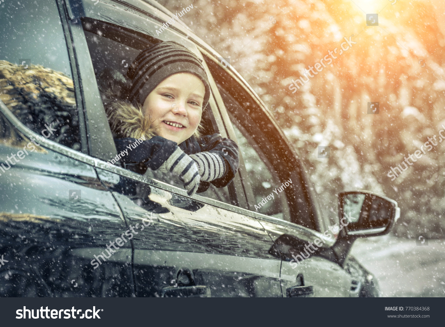 Happiness caucasian smilling boy looking out of black car window in sunny day at winter time near the forest.