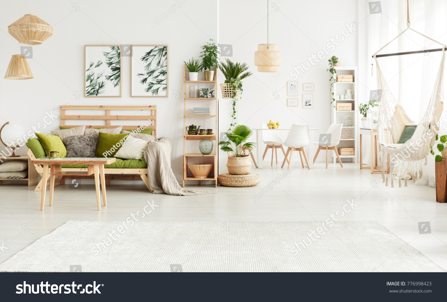 Plant on pouf and leaves posters in bright open space interior with green couch hammock and white chairs at a table