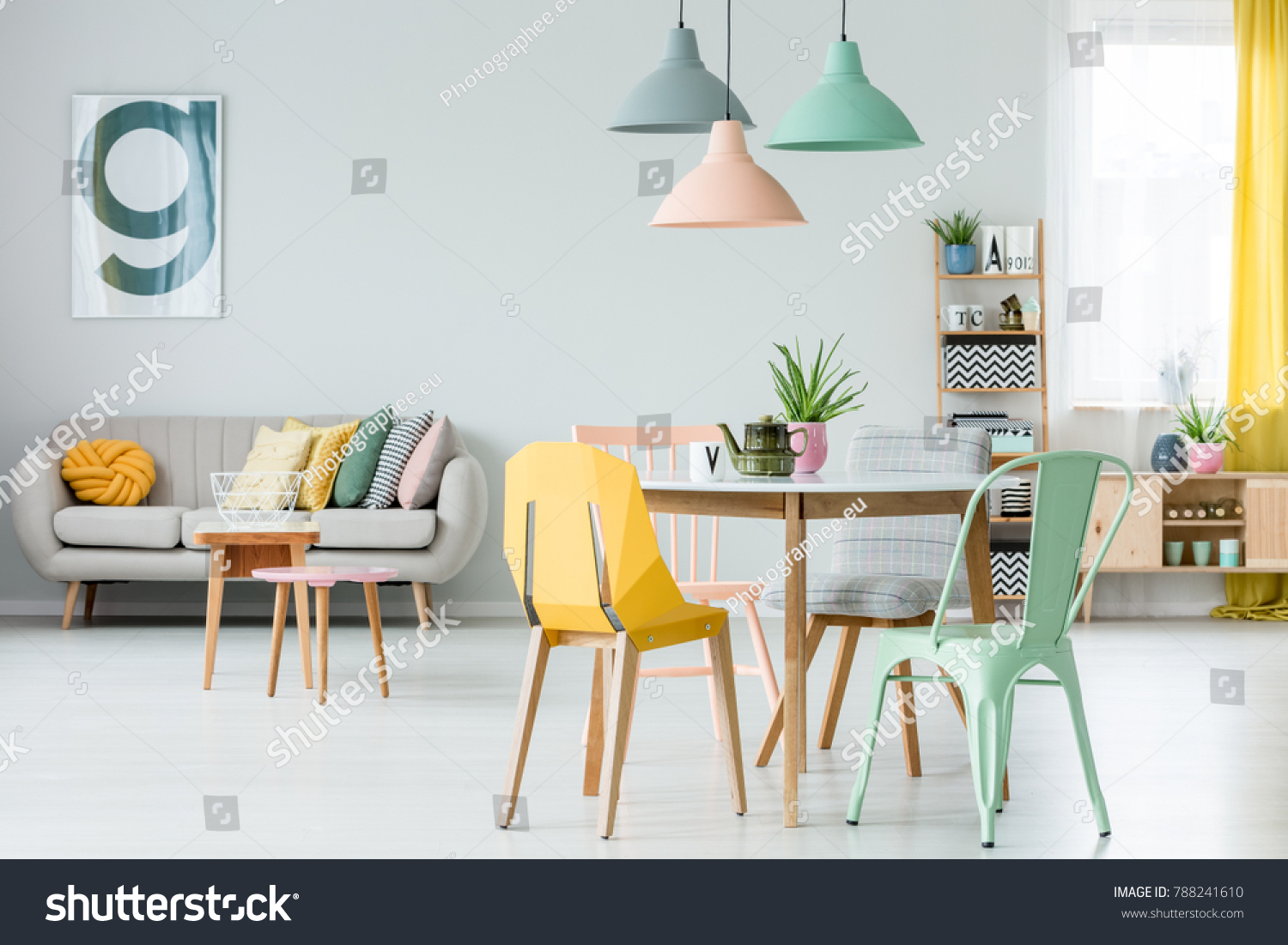Modern colorful chairs at dining table under pastel lamps in living room interior with pillows on settee against wall with poster