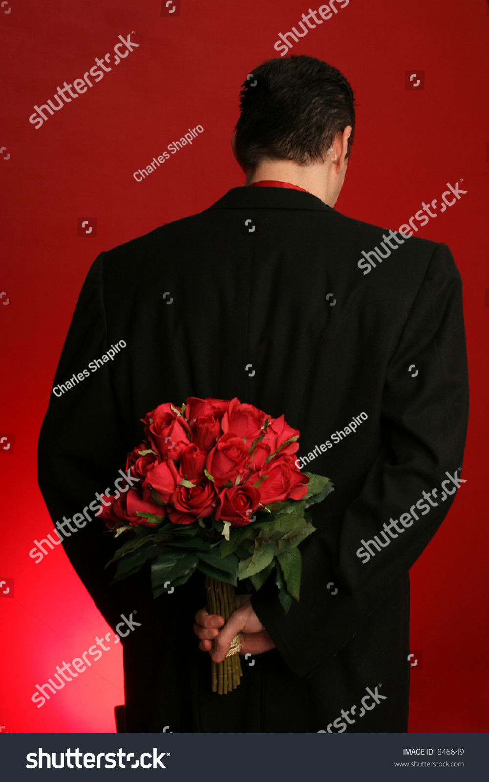 man holding out large bouquet of red roses behind his back on red backdrop