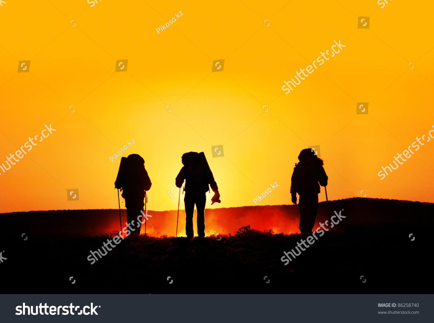 Three tourist silhouettes walking on the hills with track sticks and backpacks at sunset orange background. Free space for text good template for web design