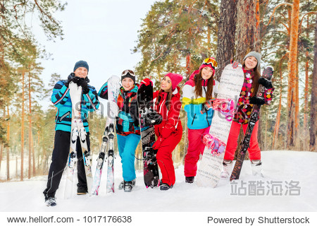The cheerful company friends  snowboarders and skiers standingsnow-covered slope in the background of forest and sky. Horizontal.Four girls and a boy in a ski suit with skis and snowboards sta