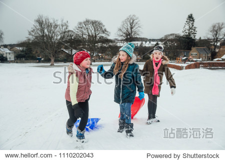 Three little girls are talking while they are out playing in the snow with sleds. 