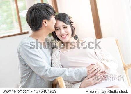 Happy excited pregnant couple having fun at home. Cheerful expectant mom sitting on couch dad kiss forehead look out window smiling laughing. Enjoying pregnancy mother’s day concept