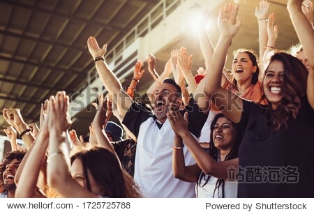 Crowd of sports fans cheering during a match in stadium. Excited people standing with their arms raised clapping and yelling to encourage their team.