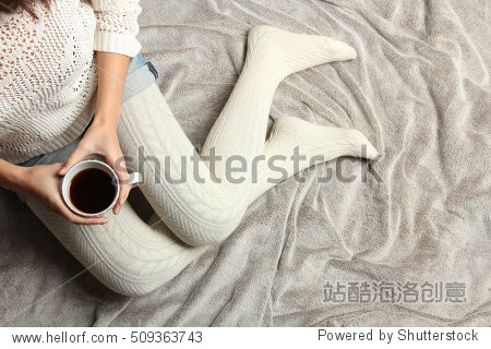 Soft cozy photo of woman in warm sweater on the bed with cup of tea in hands top view point. Composition with warm plaid cup of hot drink and female legs
