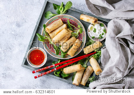 Fried spring rolls with red and white sauces served in china plate on wood tray with fresh green salad and wooden chopsticks over gray blue texture background. Flat lay space. Asian food