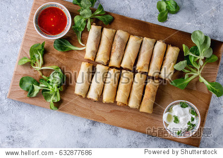 Fried spring rolls with red and white sauces served on wood serving board with fresh green salad and wooden chopsticks over gray blue texture background. Flat lay space. Asian food