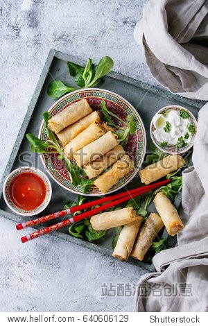 Fried spring rolls with red and white sauces served in china plate on wood tray with fresh green salad and wooden chopsticks over gray blue texture background. Flat lay space. Asian food