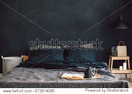 Black stylish loft bedroom. Unmade bed with breakfast and reading on tray. Lamp and interior decor over blank blackboard wall with copyspace. Cozy modern living space.