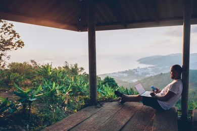 young man traveler using laptop computer while enjoying beautiful tropical sunset in thailand. freelancer guy working on freelance on netbook during vacation holidays in summer. male adventure blogger