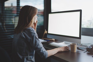 view from back to female student is talking on smartphone sitting at desk in front of monitor with blank space for design. mockup screen with copy space. woman makes a business call