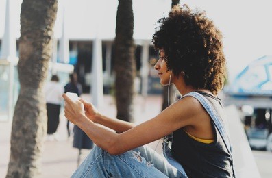 portrait of a girl with afro hair who watches a video on a mobile phone while resting outside. a young dark skinned woman uses headphones to communicate with friends via skype via modern smartphone